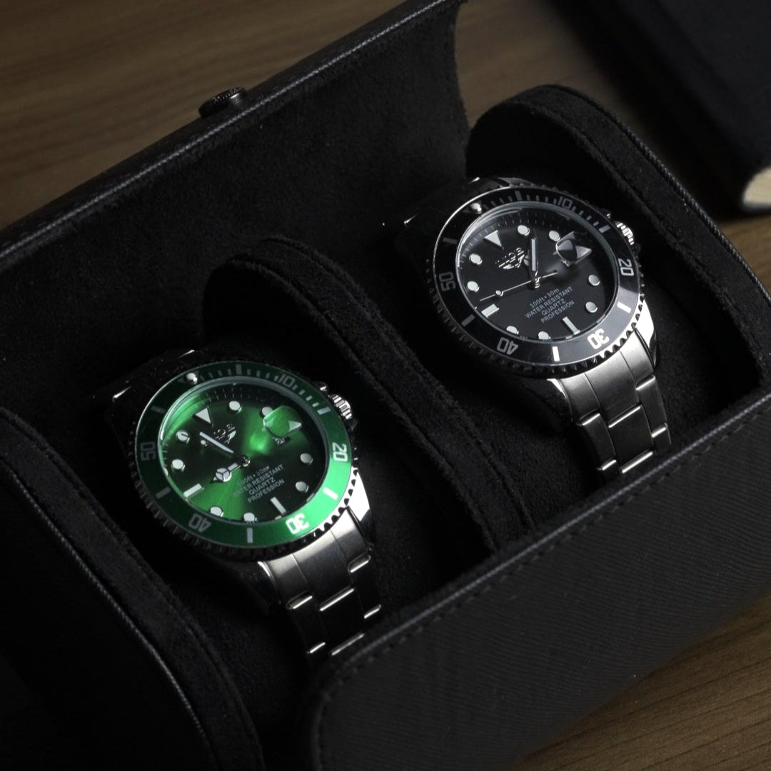 Two watches, one with a green face and silver case, the other with a black face and silver case, in a black box on a wooden surface.