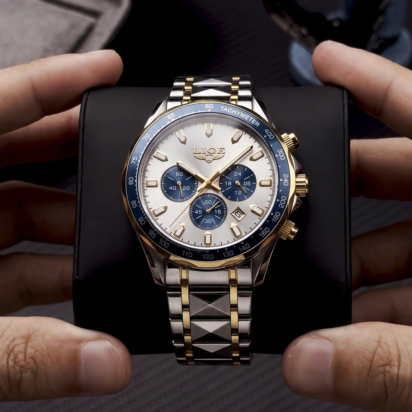 A luxury two-tone watch with a silver and gold metal bracelet, a white dial with blue subdials, and a blue bezel, held by hands on a black display block.
